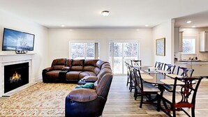 A modern living room with a leather sectional sofa, a fireplace, a dining area with a long table and chairs, and a kitchen in the background.