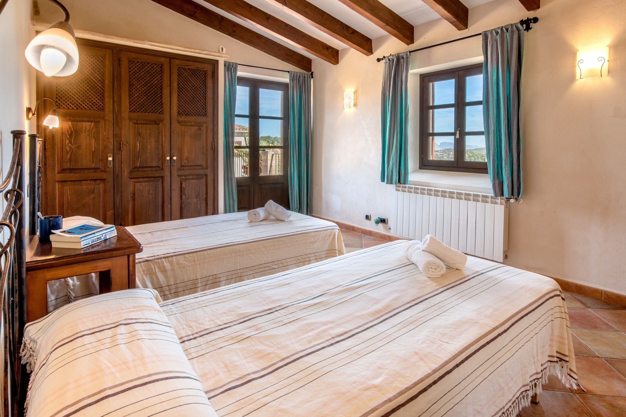 A hotel room with two beds, wooden wardrobe, a bedside table with a lamp, and a window with curtains.