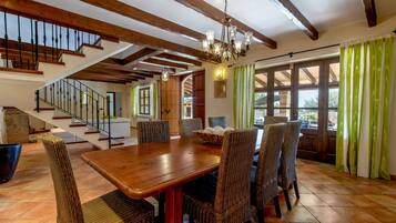 A dining area with a wooden table and wicker chairs, a staircase, and large windows with curtains.