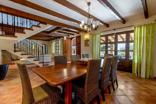 A dining area with a wooden table and wicker chairs, a staircase, and large windows with curtains.
