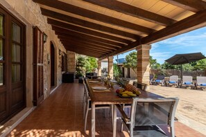 Um pátio externo com mesa de madeira posta para refeições, rodeado por cadeiras e uma área coberta com vista para o jardim.