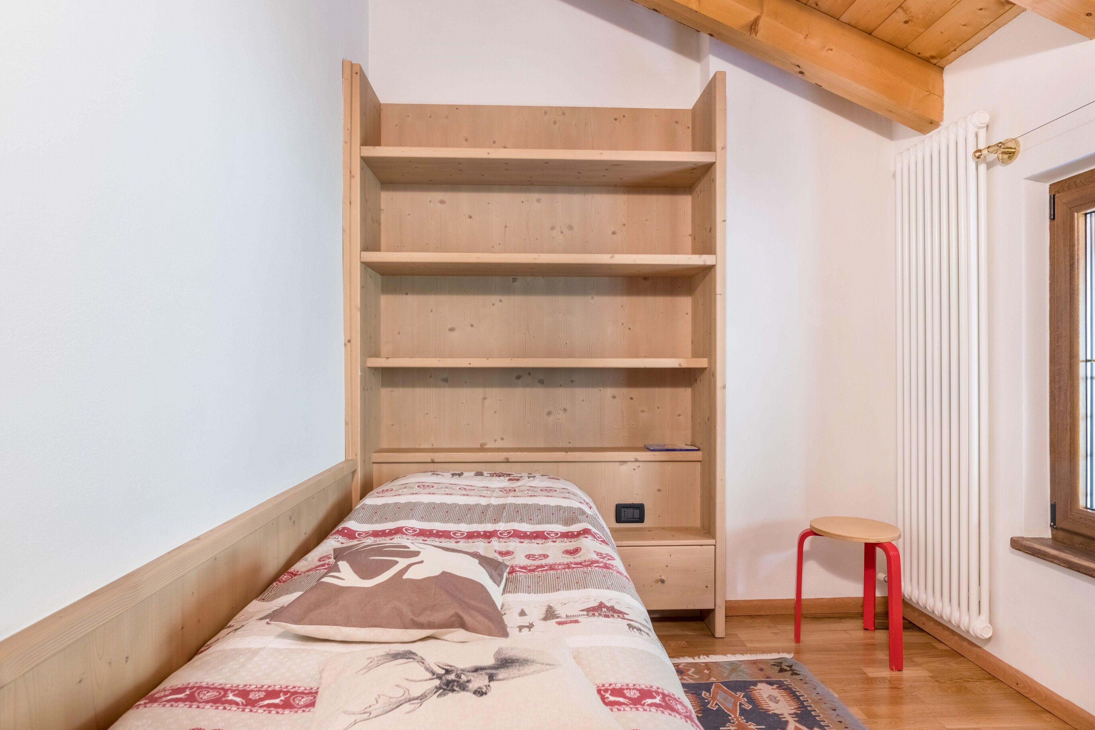 Een kamer met een bed, een houten plankensysteem, een rode kruk en een raam met een radiator.