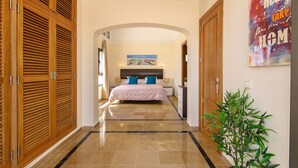 Un pasillo que conduce a un dormitorio con una cama, puertas de madera, una planta en maceta y un cuadro en la pared.