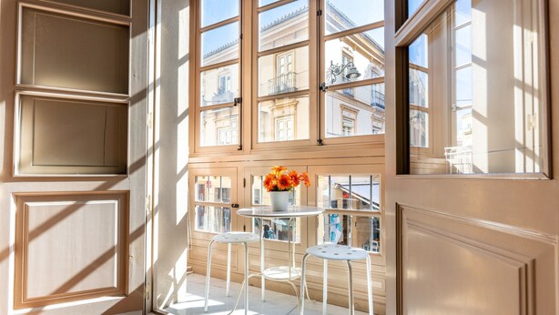 Ein Zimmer mit großem Fenster, ein Balkon mit zwei weißen Stühlen und einer Blumenvase.