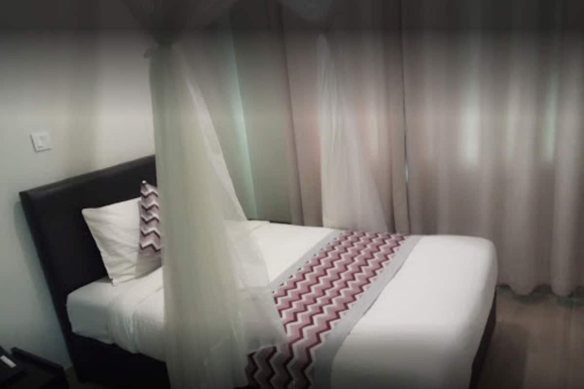 Ein Bett mit weißer Bettdecke und einer deckenartigen, zickzackmusternden Wolldecke, ein dunkles Kopfteil und transparente Vorhänge.