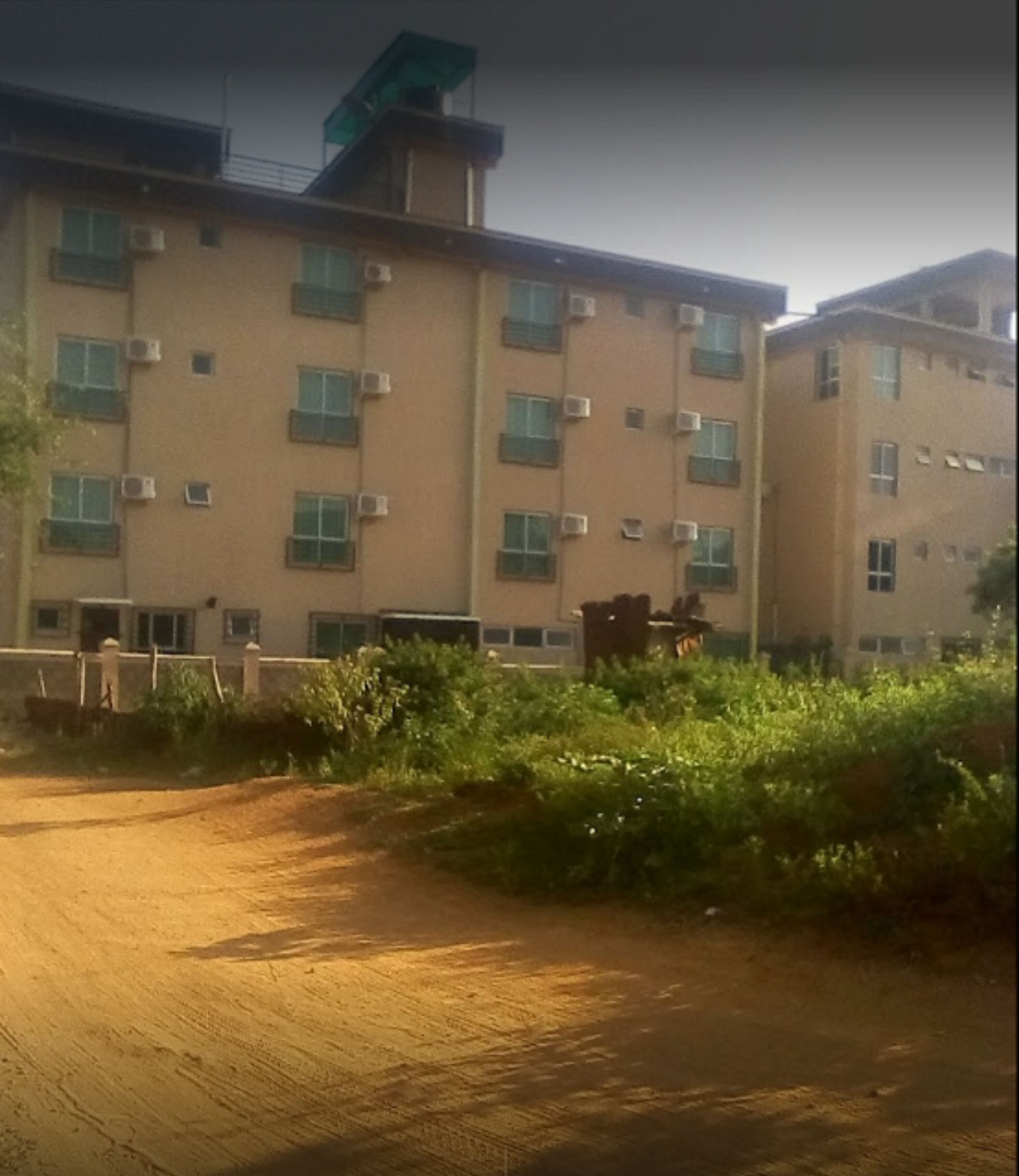 A multi-story building with a flat roof and multiple windows, surrounded by greenery and a dirt path.