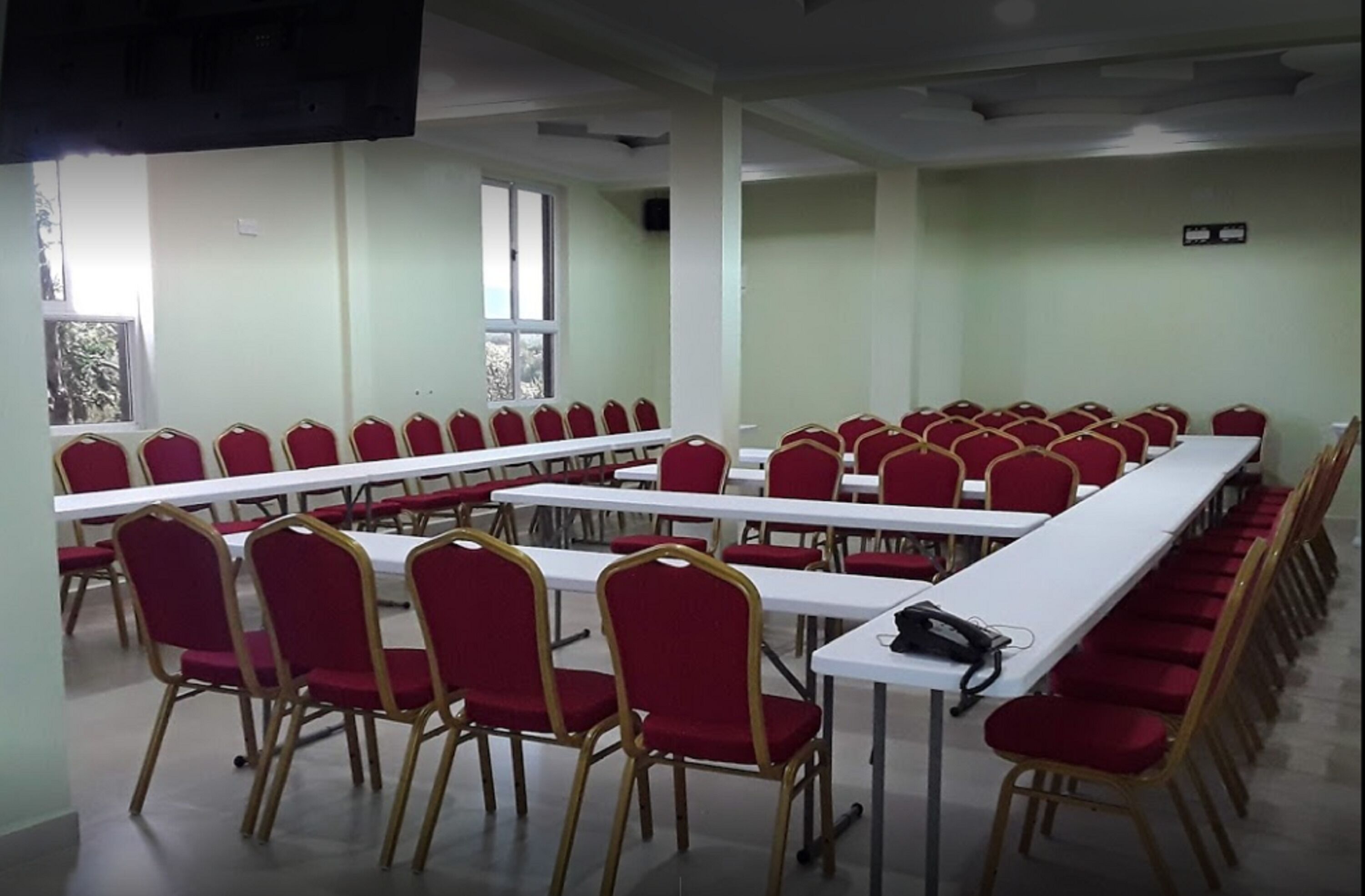 Una sala conferenze con file di sedie rosse e tavoli bianchi, ampie finestre e un televisore a schermo piatto montato a parete.