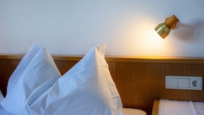 A hotel room with a bed, white pillows, a wooden headboard, a wall-mounted light, and a power outlet.