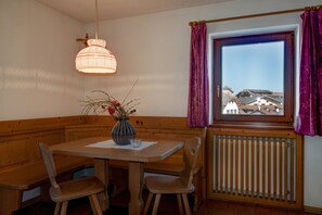 Un coin repas avec une table et des chaises en bois, un vase avec des branches, un luminaire suspendu, une fenêtre avec des rideaux violets et une vue sur les bâtiments à l’extérieur.