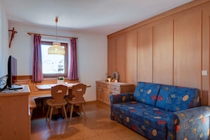 A cozy room with a wooden dining table, two chairs, a sofa, a television, and a window with curtains.