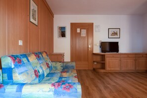 Une pièce avec un plancher en bois, un canapé à motifs floraux, un téléviseur posé sur un meuble en bois et une porte sur laquelle est affichée une annonce.