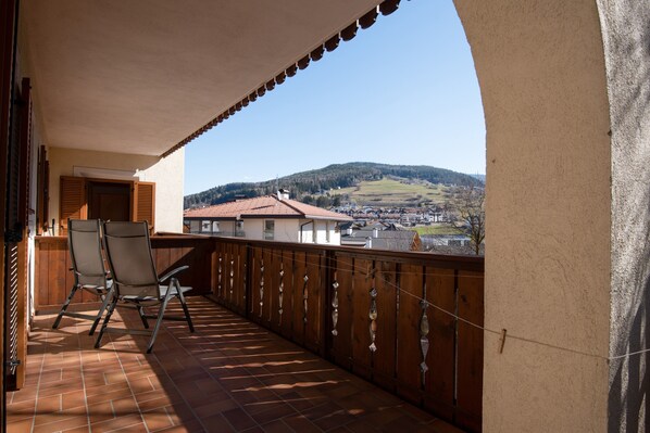 Un balcon avec deux chaises, une vue sur un paysage vallonné et, au loin, un bâtiment avec un toit rouge.