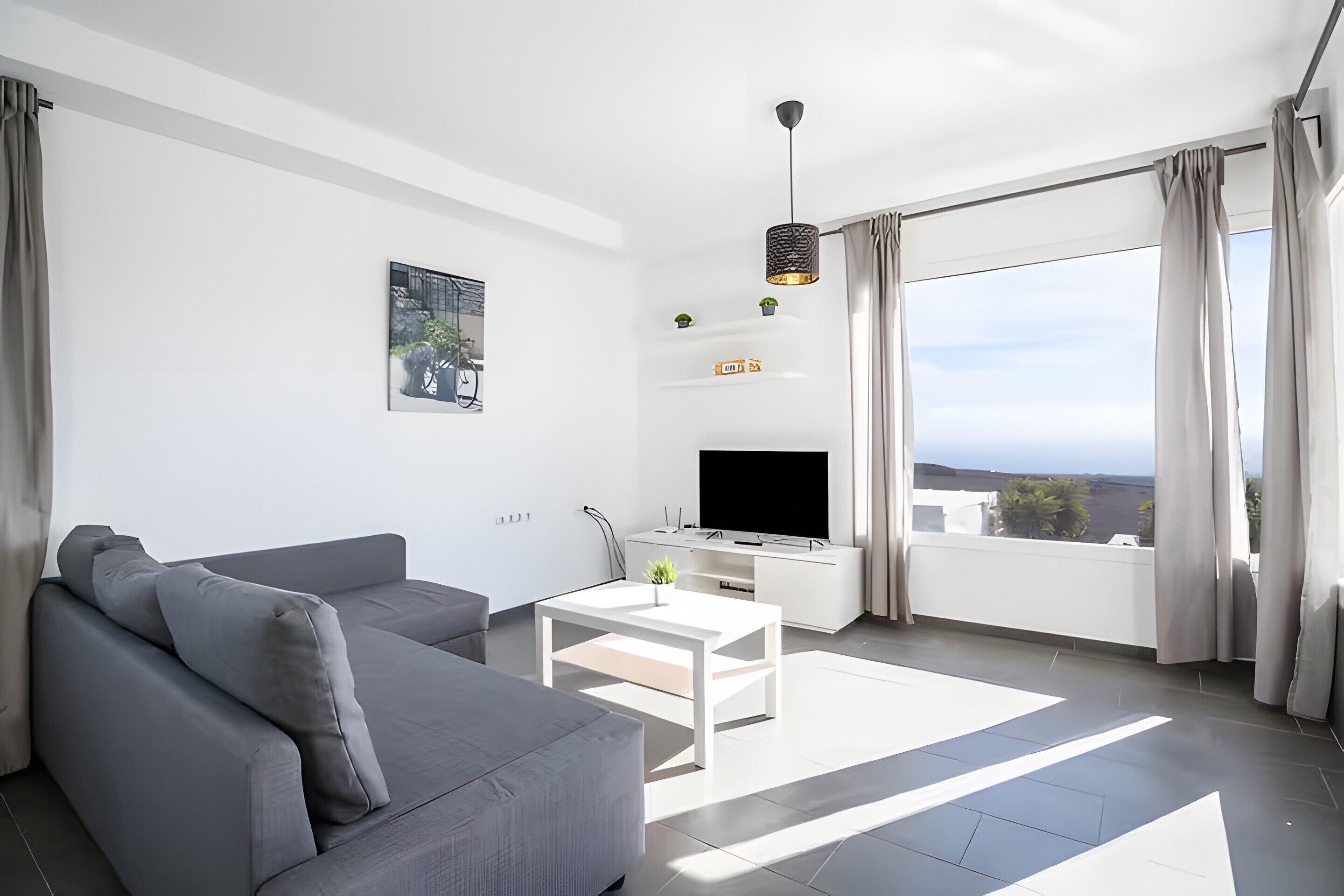 A modern living room with a grey sofa, a white coffee table, a flat-screen TV, and a balcony with curtains.