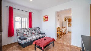 A living room with a sofa, a table, a red curtain, a window, and a kitchen area in the background. - Rural Home "La Ermita" with Pool, Terrace, Wi-Fi & Air Conditioning (Conil de la Frontera)
