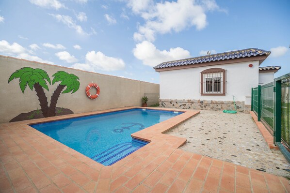 Une piscine avec une terrasse carrelée, une bouée de sauvetage, une fresque représentant des palmiers et un petit bâtiment doté d’un toit en tuiles.