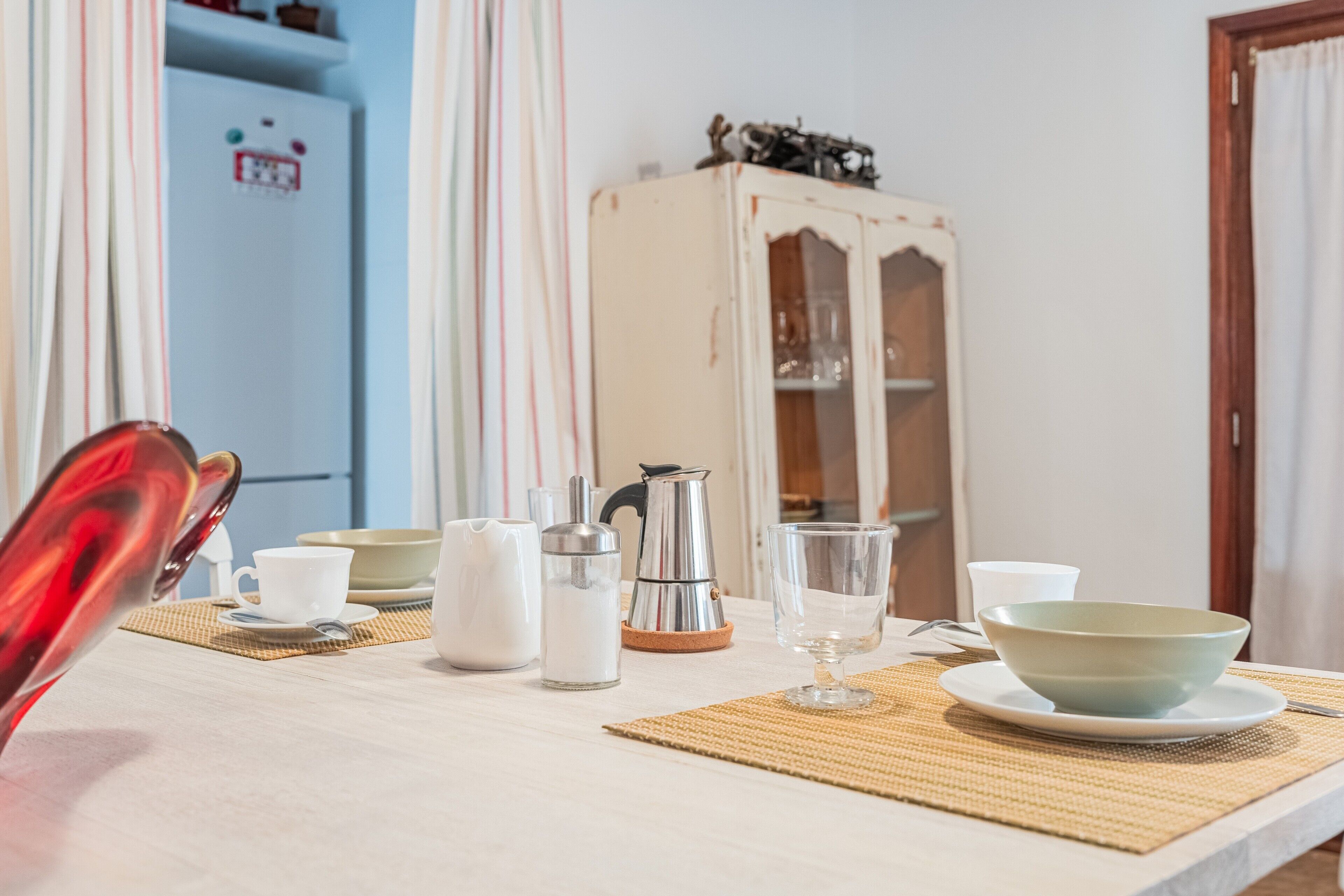 Une table à manger recouverte d’une nappe blanche, une cafetière, des tasses et un bol.