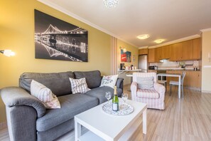 Un salon avec un canapé gris, une table basse blanche sur laquelle se trouvent une bouteille et des verres, un fauteuil rose, et une cuisine en arrière-plan.