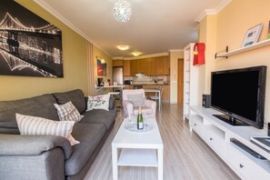 Un salon moderne avec un canapé, une table basse sur laquelle se trouve une bouteille, un téléviseur et, au fond, une cuisine.