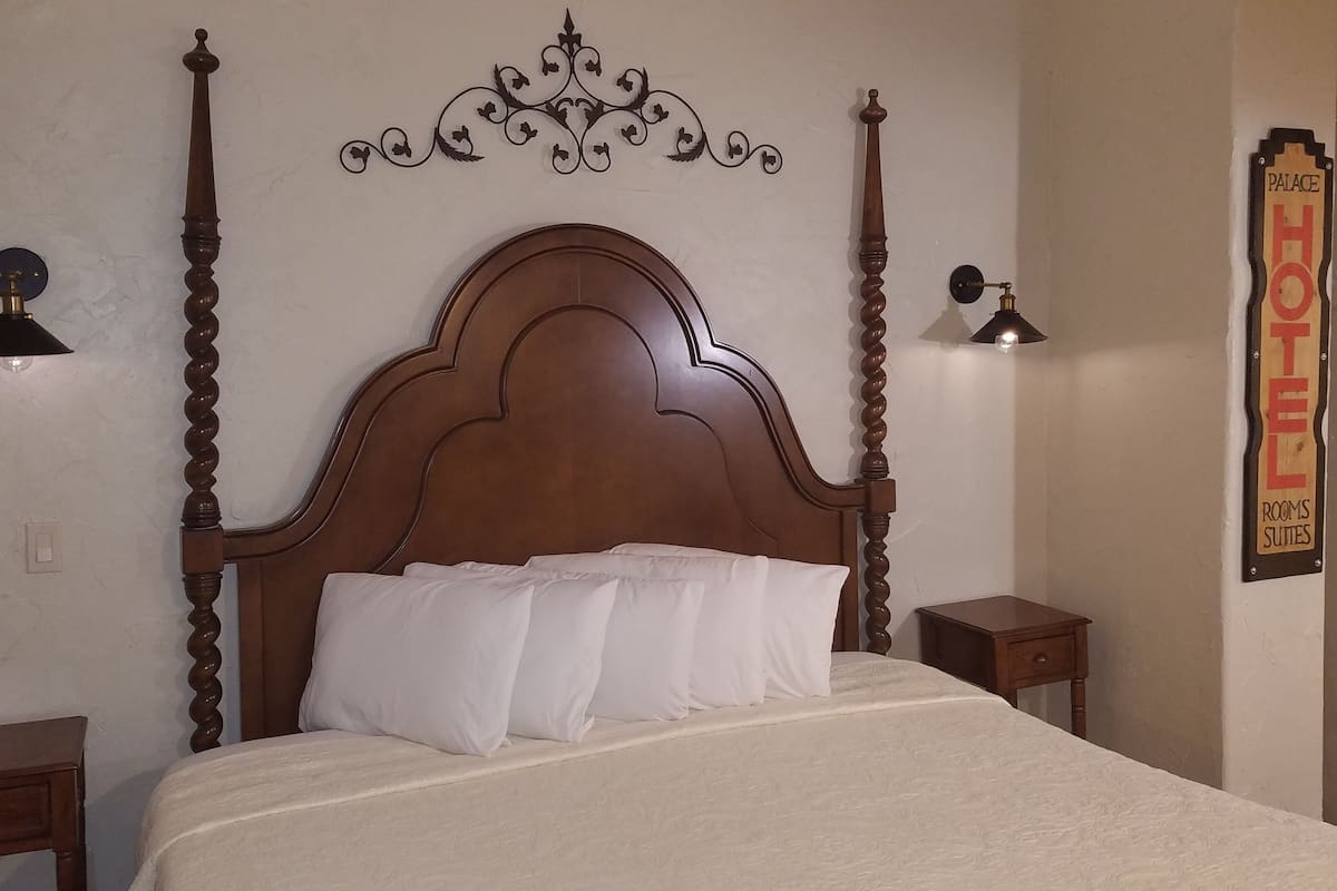 A neatly made bed with a wooden headboard, two bedside tables, and a wall-mounted sign indicating "PALACE HOTEL ROOMS SUITES".
