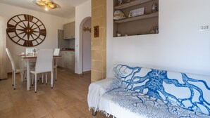 Zona pranzo con tavolo e sedie, un divano con una coperta decorativa, una sveglia a parete e mensole con oggetti ornamentali.