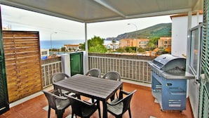 A balcony with a table and chairs, a barbecue grill, and a view of the sea and buildings.