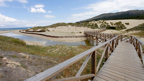 A wooden boardwalk leading to a beach with dunes and a hill in the background.