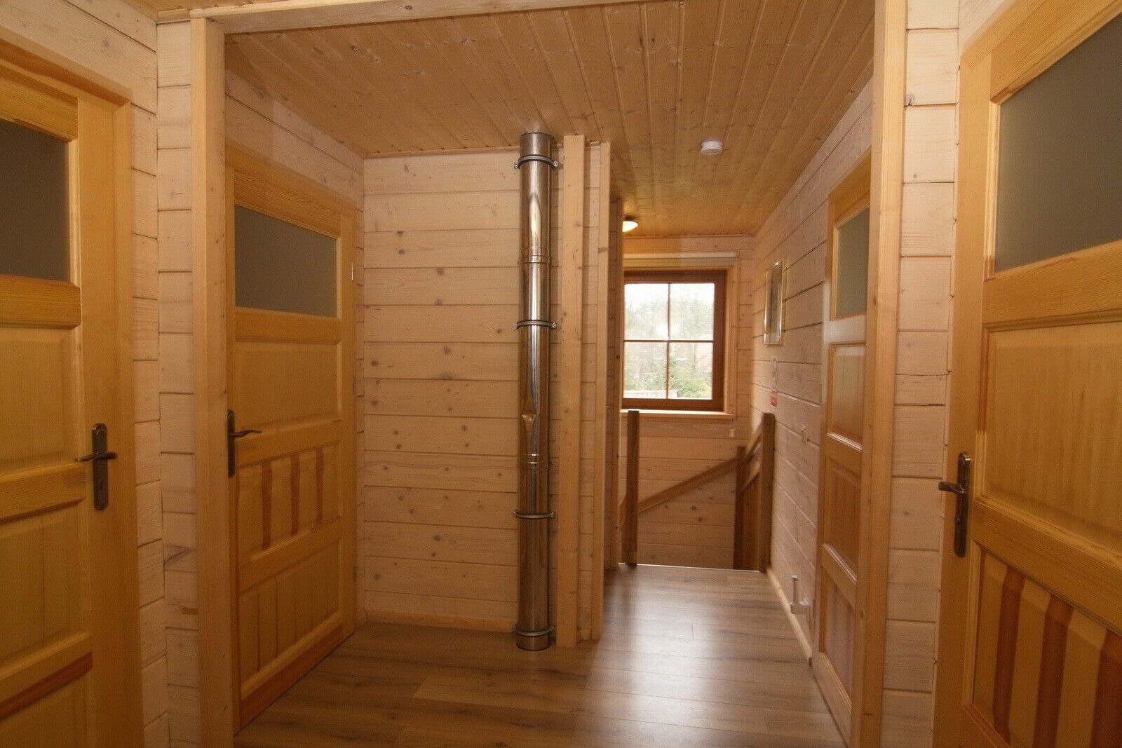 Un pasillo interior de madera con puertas, un conducto vertical en el centro y una ventana al final.
