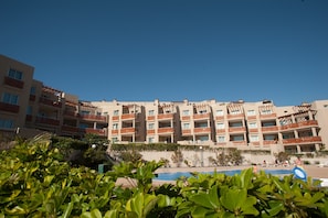 Un hotel a più piani con balconi, una piscina e una vegetazione rigogliosa in primo piano.