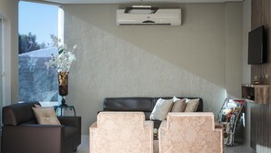 Una sala de estar con un sofá, dos sillones, una mesita de centro, un florero con flores, un aire acondicionado de pared y un mueble para revistas.