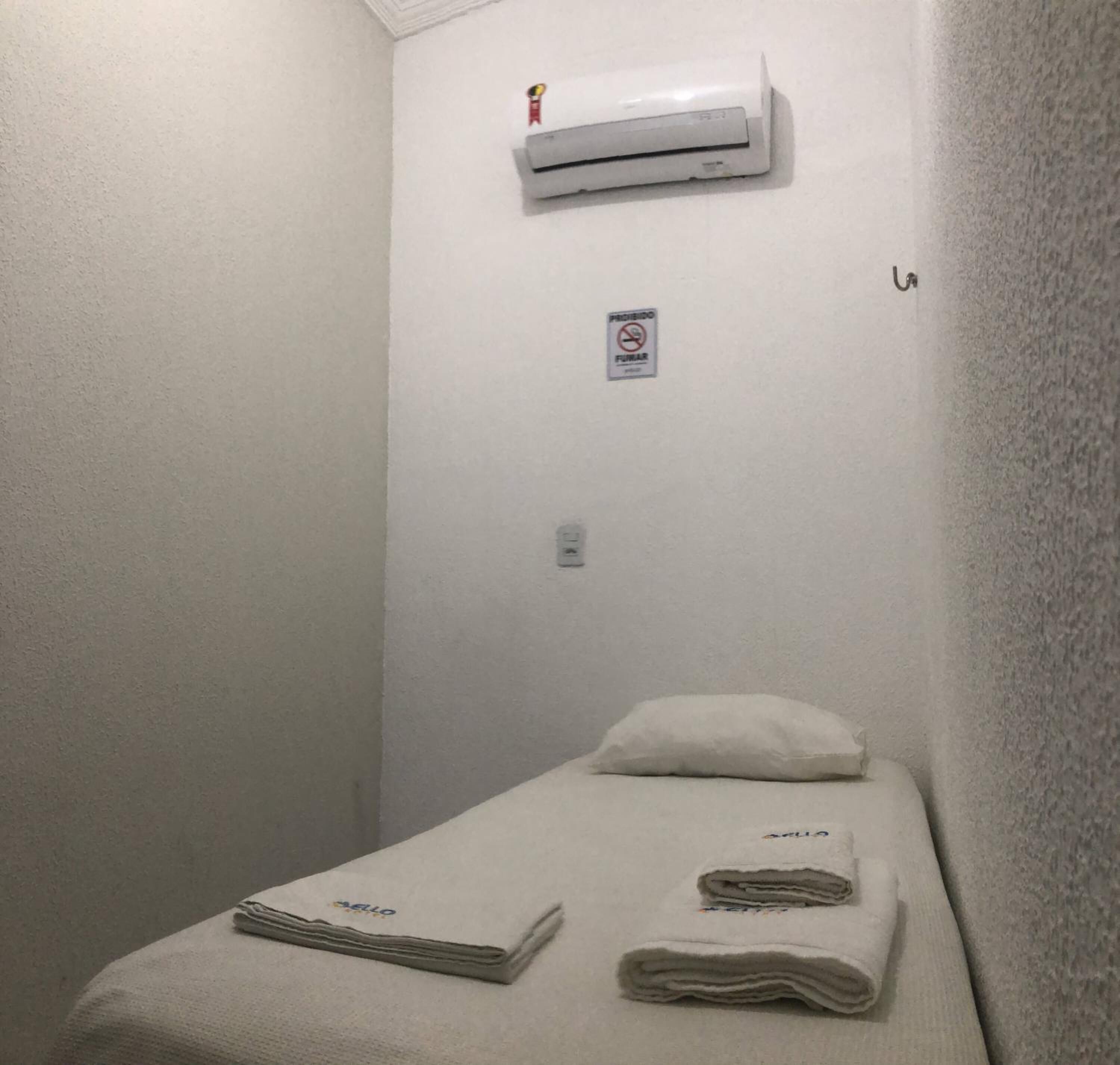 Una habitación pequeña con cama individual, pared blanca, unidad de aire acondicionado sobre la cama, letrero de 'no fumar' y una toalla doblada sobre la cama.