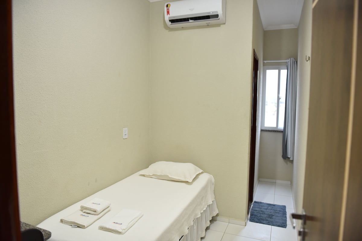 Una habitación pequeña con cama individual, colcha blanca, almohada blanca y dos toallas dobladas. Se observa una ventana con cortinas, una puerta y un aire acondicionado de pared.