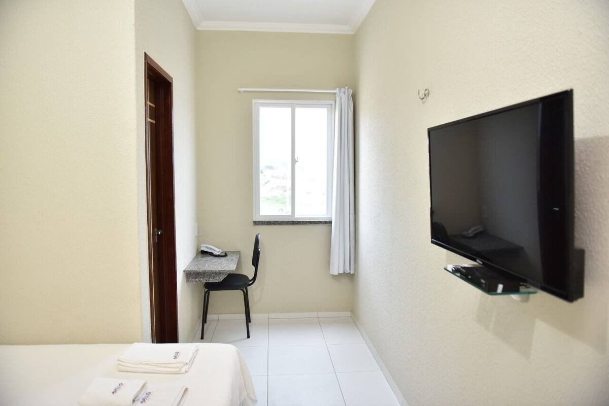 Una habitación pequeña con una cama, un escritorio con silla, una ventana con cortinas y un televisor montado.