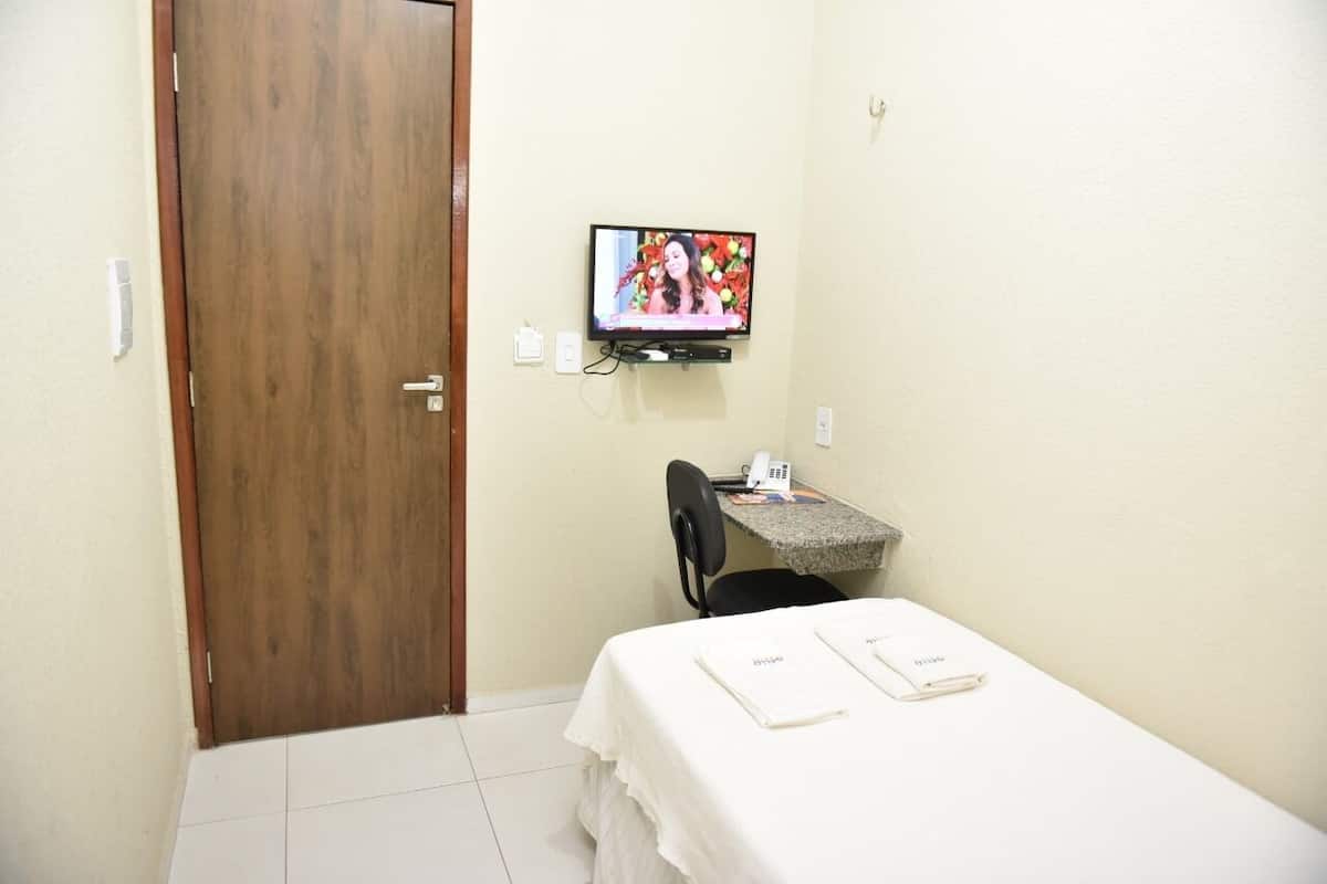 Una habitación de hotel pequeña con una puerta de madera, un televisor montado en la pared, un escritorio pequeño con una silla y una cama con una colcha blanca y dos etiquetas con nombres.