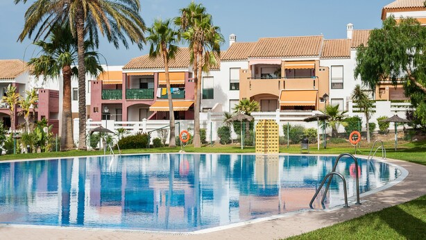 Una piscina circondata da edifici a più piani con balconi, palme e cielo sereno.