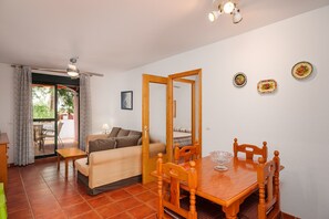 Uma área de refeições com uma mesa de madeira, cadeiras e uma tigela de vidro. Há um sofá e uma porta que dá para uma área externa.