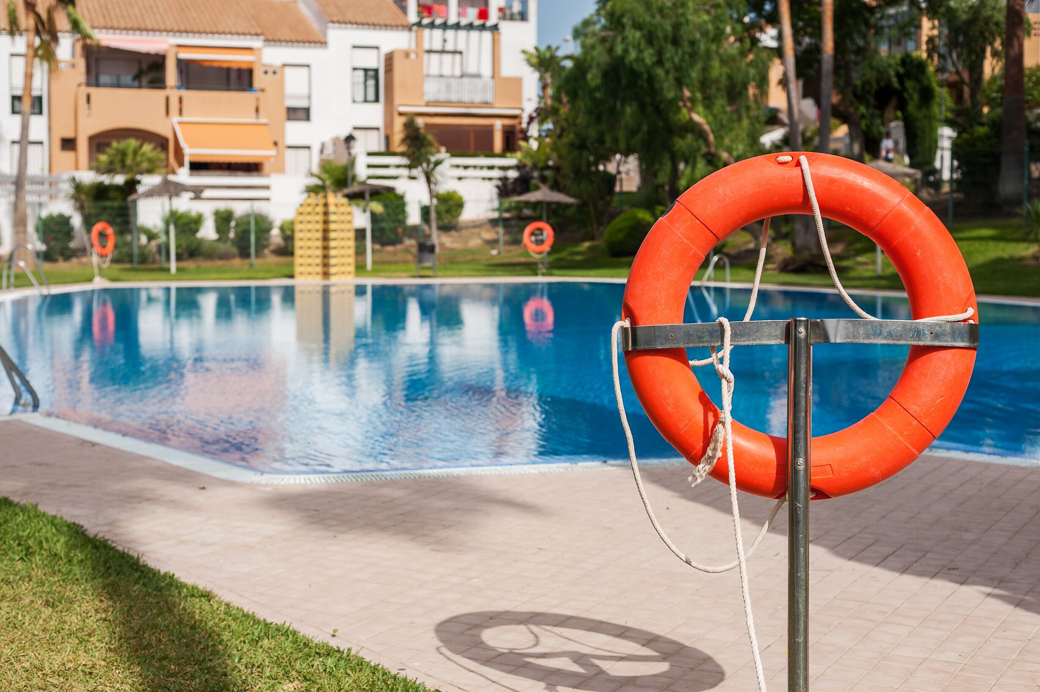 Una piscina con un flotador de salvamento sujeto a un poste, rodeada de un área pavimentada y edificios al fondo.