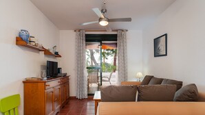 Una stanza con un letto, una TV, un armadio in legno, una sedia verde, una ventola a soffitto e un balcone con tavolo e sedie.