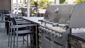 A stainless steel grill with multiple burners and a side cabinet, surrounded by outdoor seating with metal chairs and a stone wall backdrop.