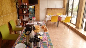 A dining area with a long table set for a meal, including plates, cutlery, and a variety of food items. There are chairs around the table, and a refrigerator with beverages is visible in the background. - Complexe Hôtelier Asthoria palace (Abidjan)