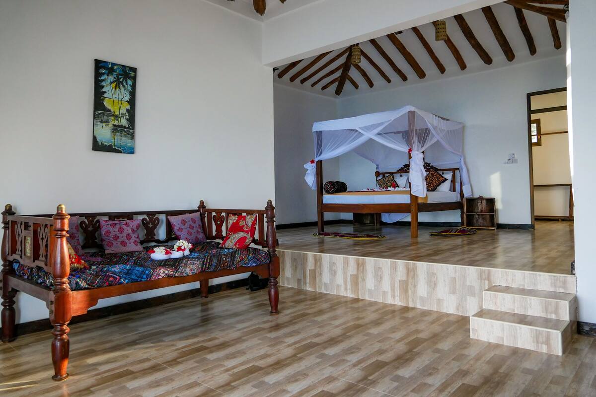 A room with a wooden sofa, a bed with a canopy, a painting on the wall, and a staircase.