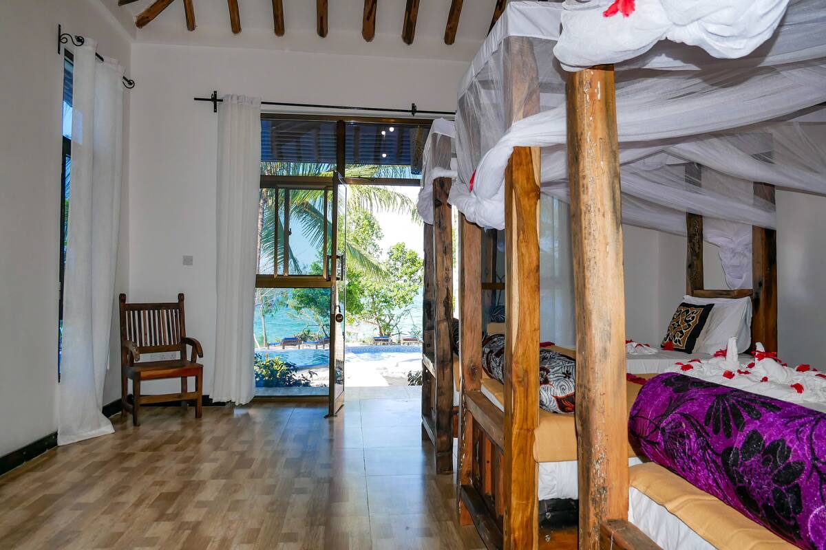 A room with a wooden floor, a bed with a purple and white patterned bedspread, a wooden chair, and a view of a pool and palm trees through a glass door.