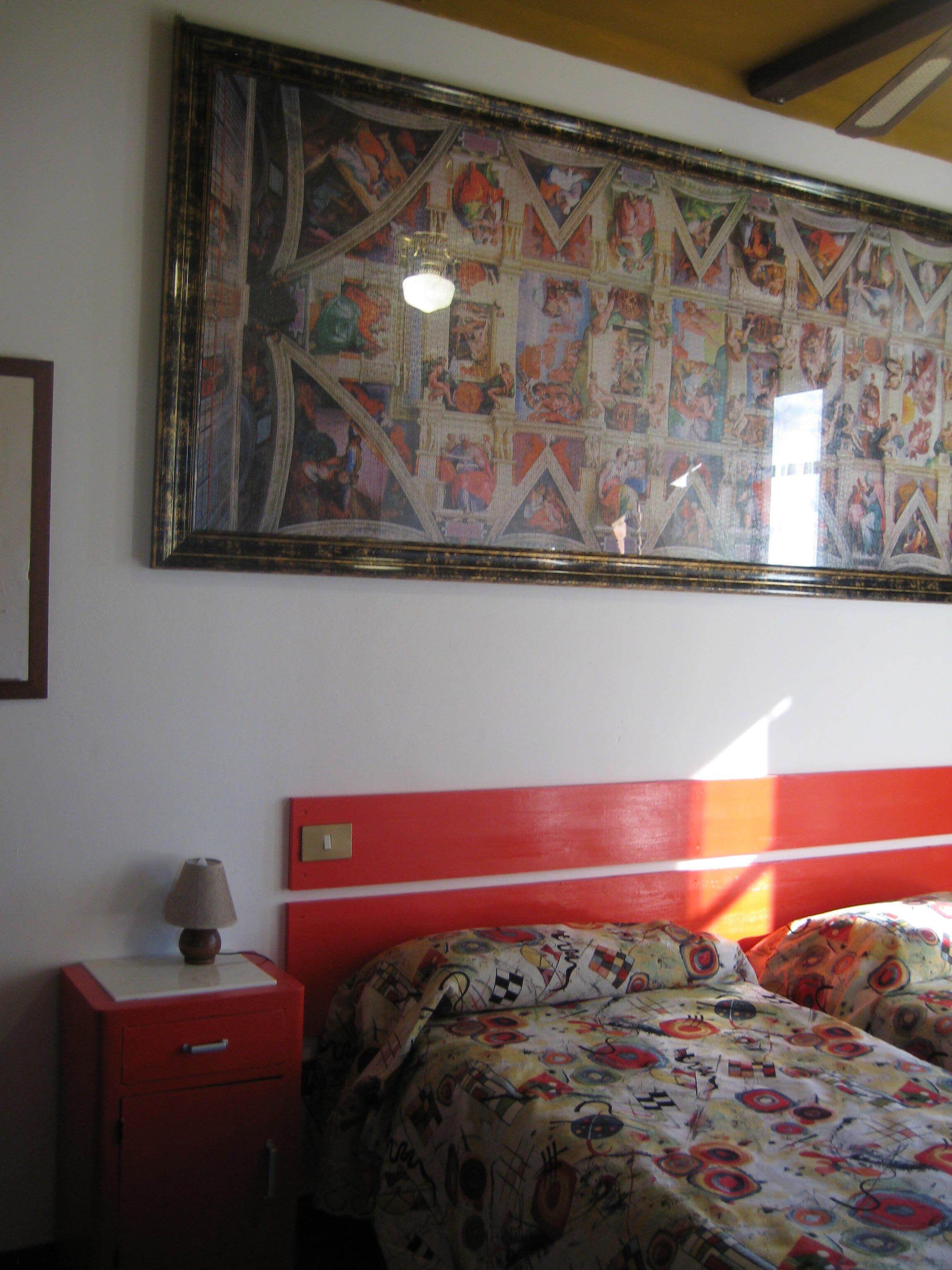 A room with a large wall mural, a red bed, a bedside table with a lamp, and a patterned bedspread.