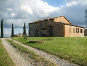 Un bâtiment rustique en pierre, auquel mène un chemin de gravier, entouré de pelouse et de deux grands arbres.