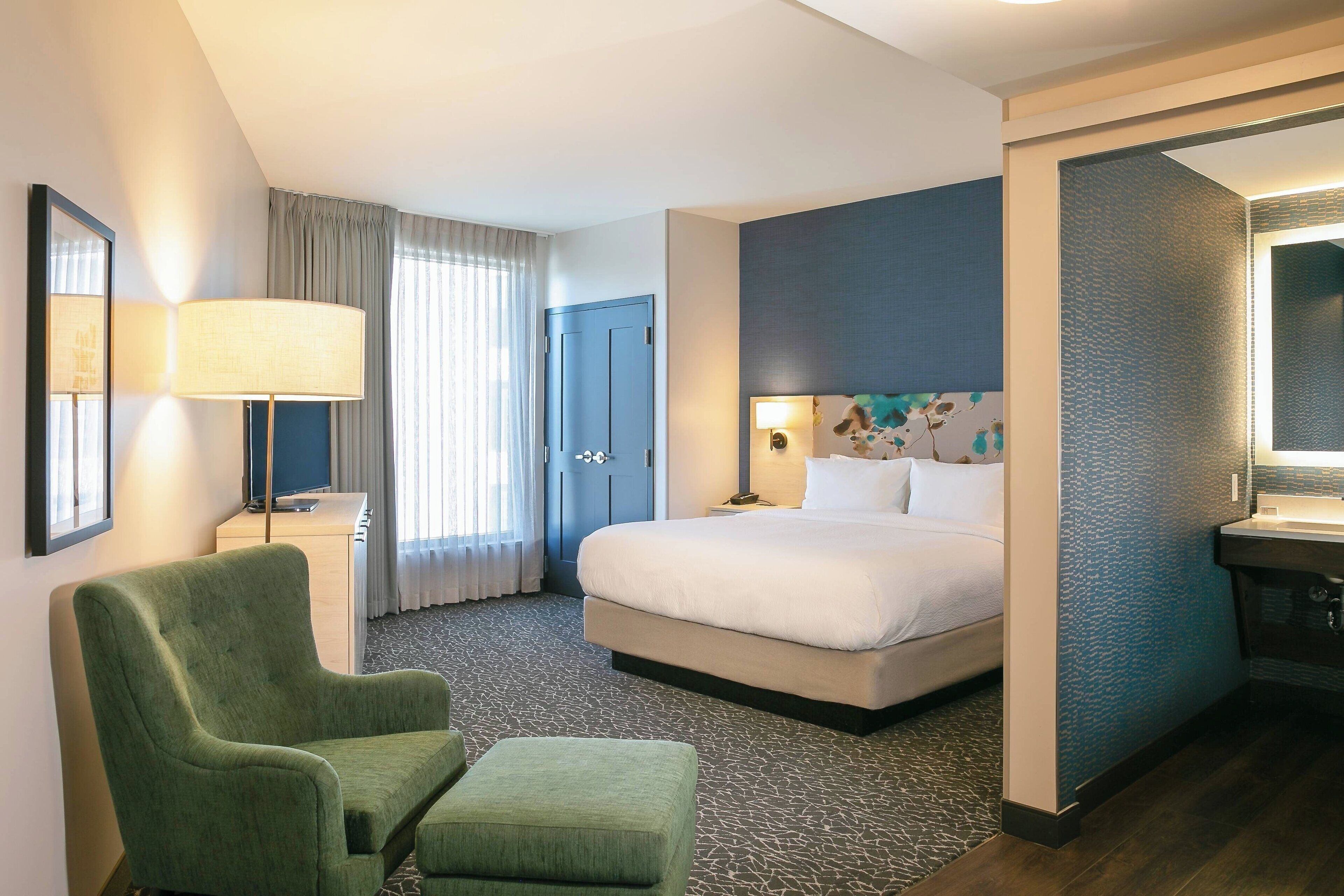 Une chambre d’hôtel avec un lit, un fauteuil vert, un petit pouf, un bureau avec une lampe et une salle de bain avec un miroir et un lavabo.