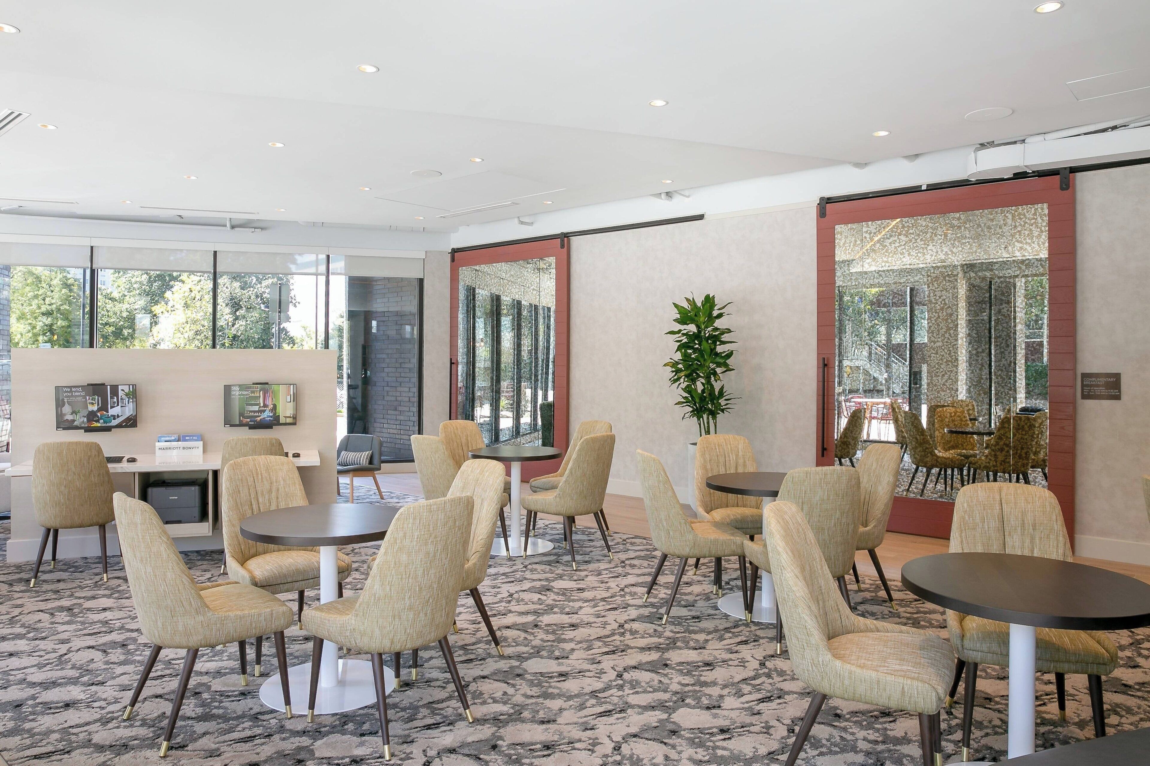 A modern hotel lobby with round tables and beige upholstered chairs, large windows, and a carpeted floor.