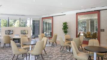 A modern hotel lobby with round tables and beige upholstered chairs, large windows, and a carpeted floor.