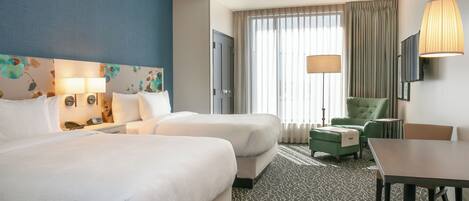 A hotel room with two beds, a desk, a chair, a table, a TV, and a large window with curtains.