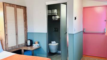 A room with a bed, a small table with a kettle and cups, a pink door, and a toilet visible through an open door.