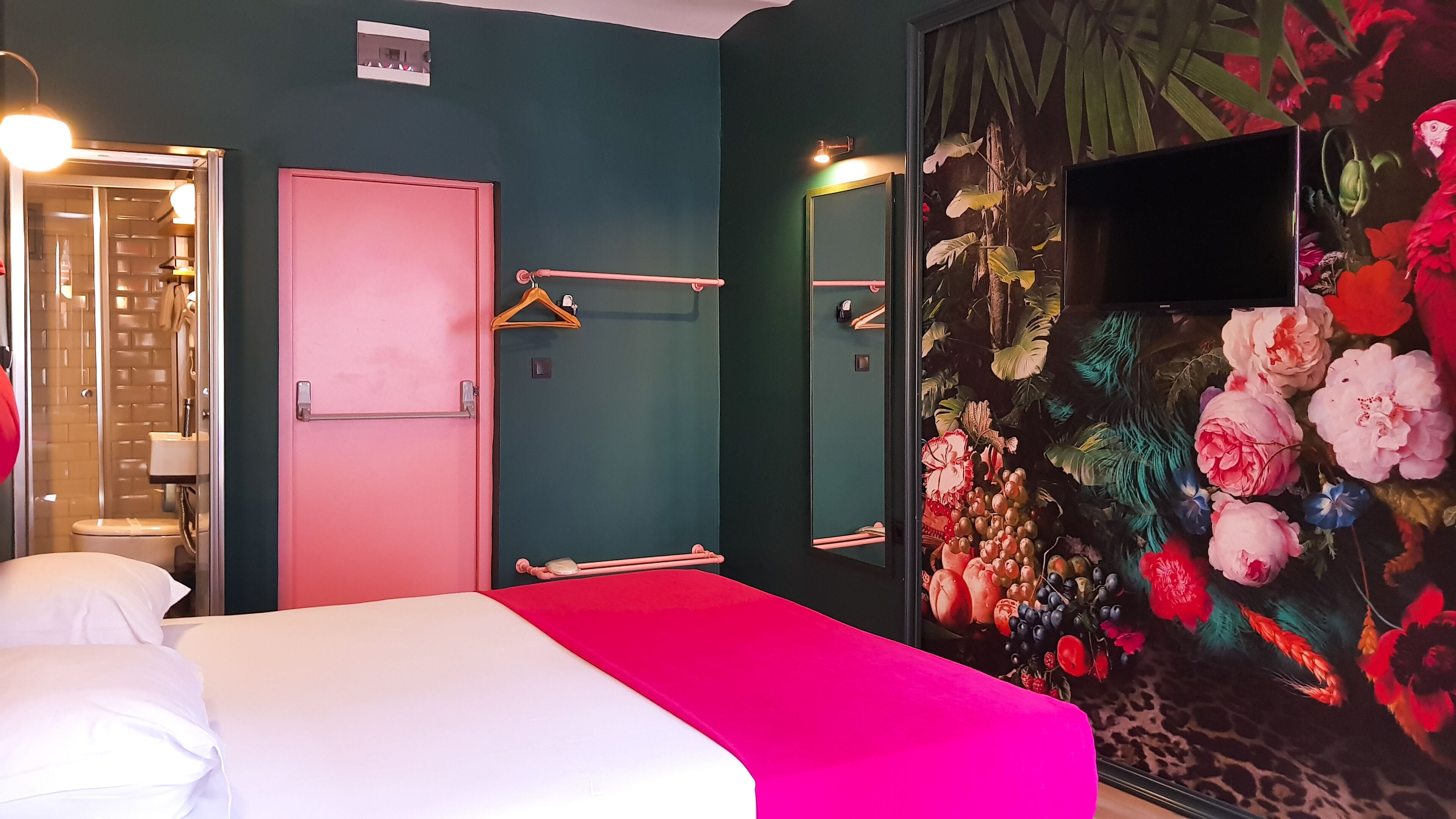 A room with a bed, a pink door, a mirror, and a wall with a floral mural.