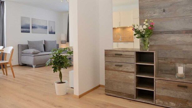 A modern living room with a grey sofa, a dining table with chairs, a potted plant, a wooden cabinet, and a kitchen area visible through an open door.